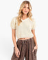 A woman modeling a cream-colored short-sleeve ribbed knit crop top with a V-neck, paired with brown drawstring pants, against a plain white studio background.