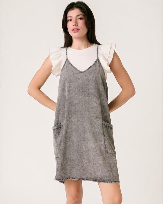 Woman wearing a gray denim pinafore dress with white ruffled sleeves on a white background