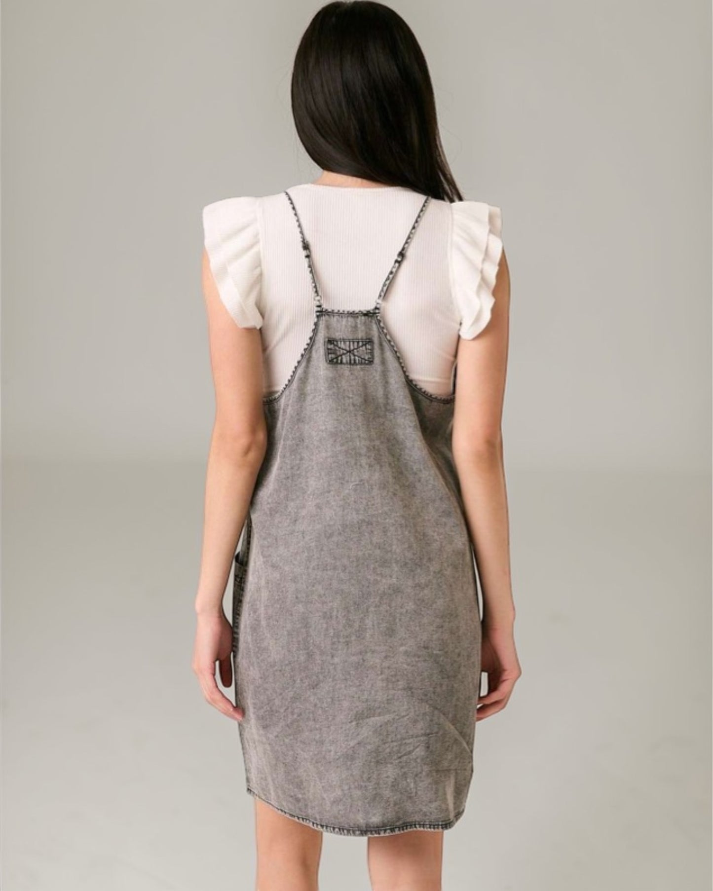 Person wearing a gray denim pinafore dress over a white blouse on a plain background