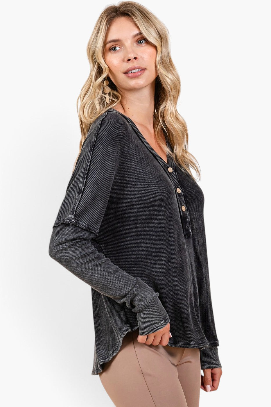 A woman wearing a dark charcoal gray washed long-sleeve henley top with button details, paired with beige pants, photographed against a clean white studio background.