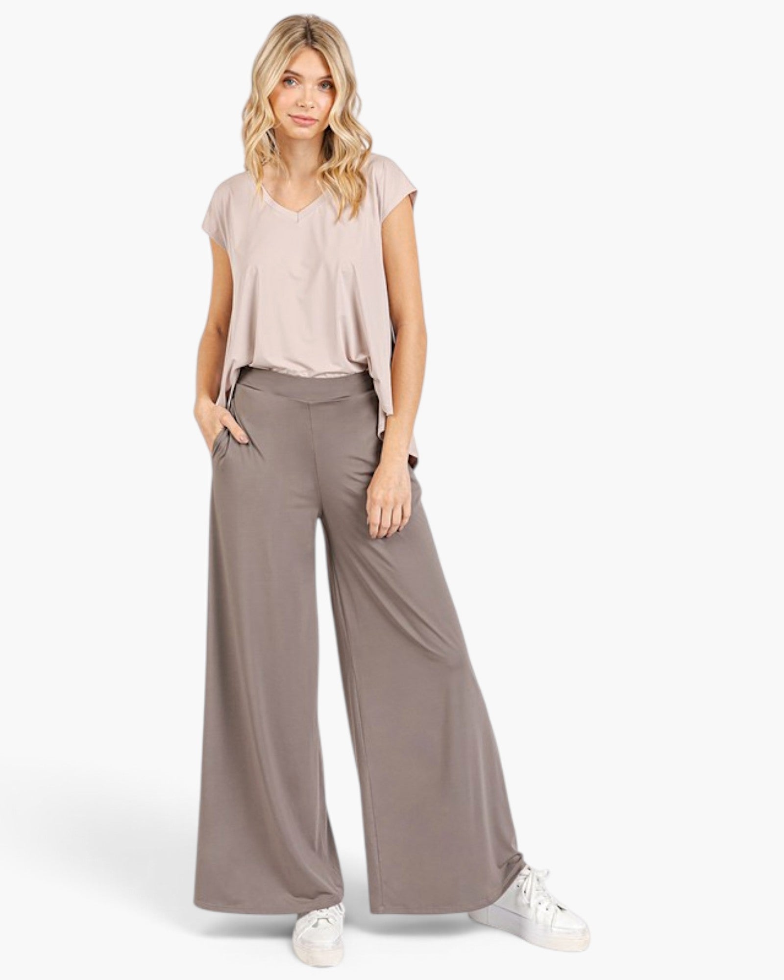 A woman modeling a light beige short-sleeve V-neck top and taupe wide-leg pants with white sneakers, set against a plain light gray studio background.
