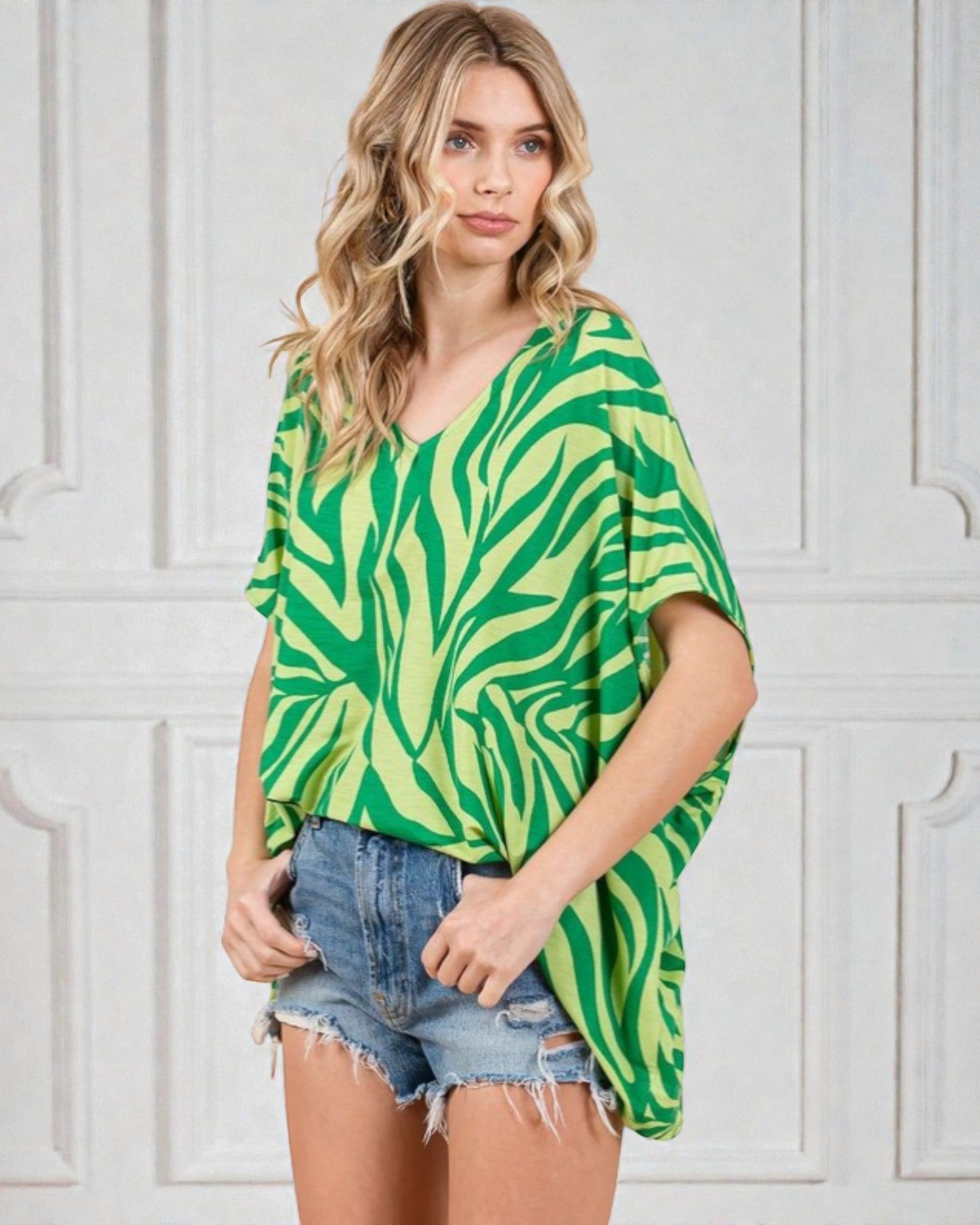 Woman wearing a green and yellow patterned top with denim shorts against a white wall.