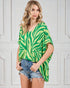 Woman wearing a green and yellow patterned top with denim shorts against a white wall.
