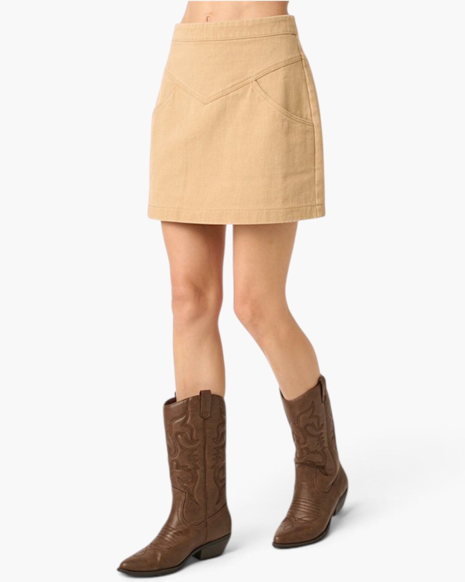 A beige mini skirt with a V-shaped waistband and side pockets, shown on a model wearing brown western boots against a light gray background.