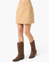 A beige mini skirt with a V-shaped waistband and side pockets, shown on a model wearing brown western boots against a light gray background.