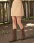 Person wearing a beige skirt and brown cowboy boots standing on a path with wooden railings in the background.