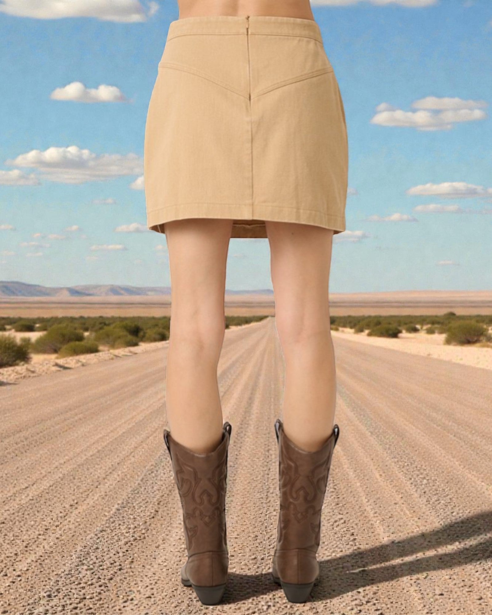Person wearing a beige skirt and brown boots standing on a desert road with a clear blue sky.
