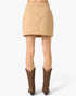 A tan mini skirt shown from the back on a model, paired with brown cowboy boots against a light gray studio background.