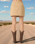 Person wearing a beige skirt and brown boots standing on a desert road with a clear blue sky.