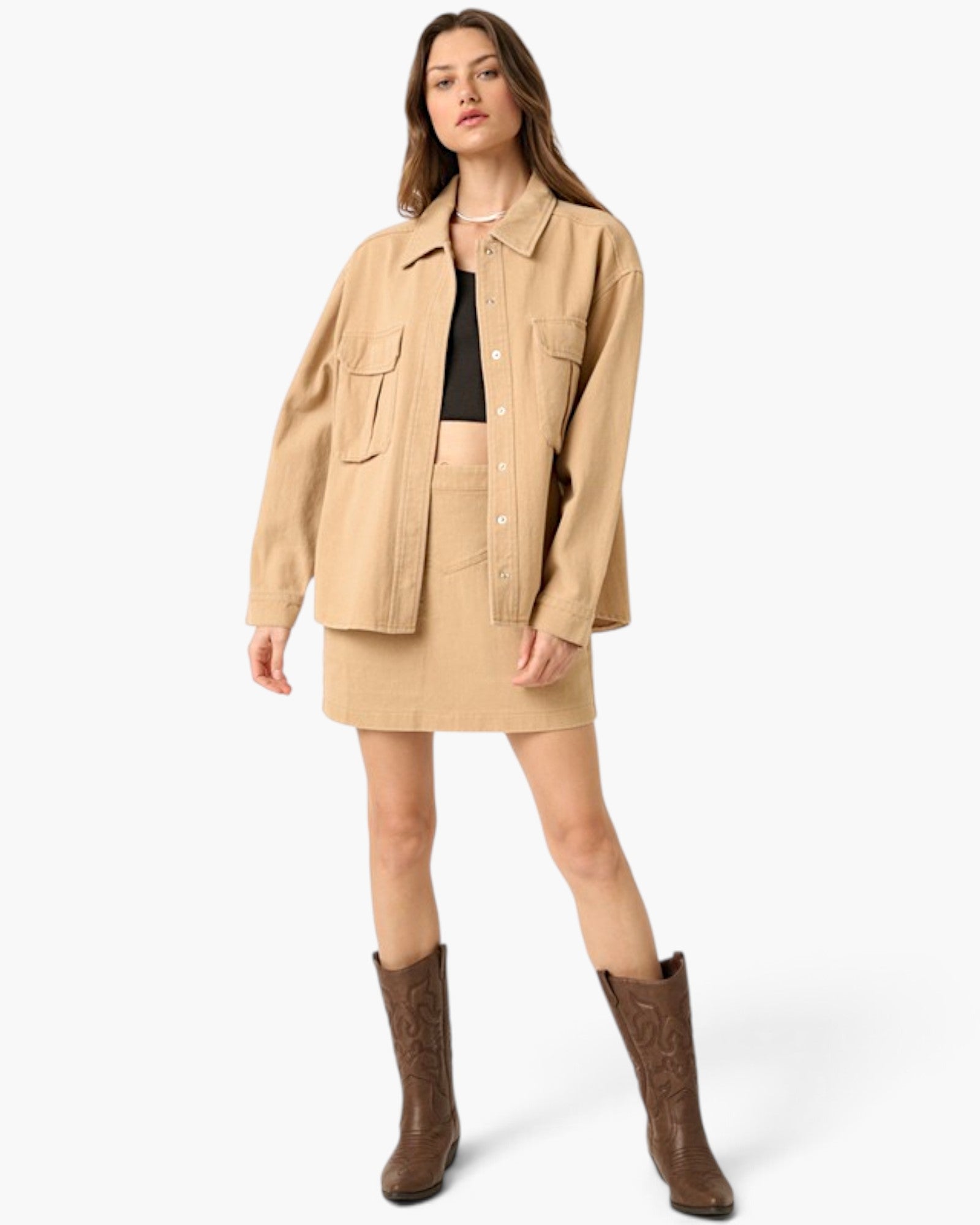 A model stands against a light gray studio background wearing a beige button-front overshirt with flap chest pockets, a matching beige mini skirt, a black cropped top, and brown cowboy boots.