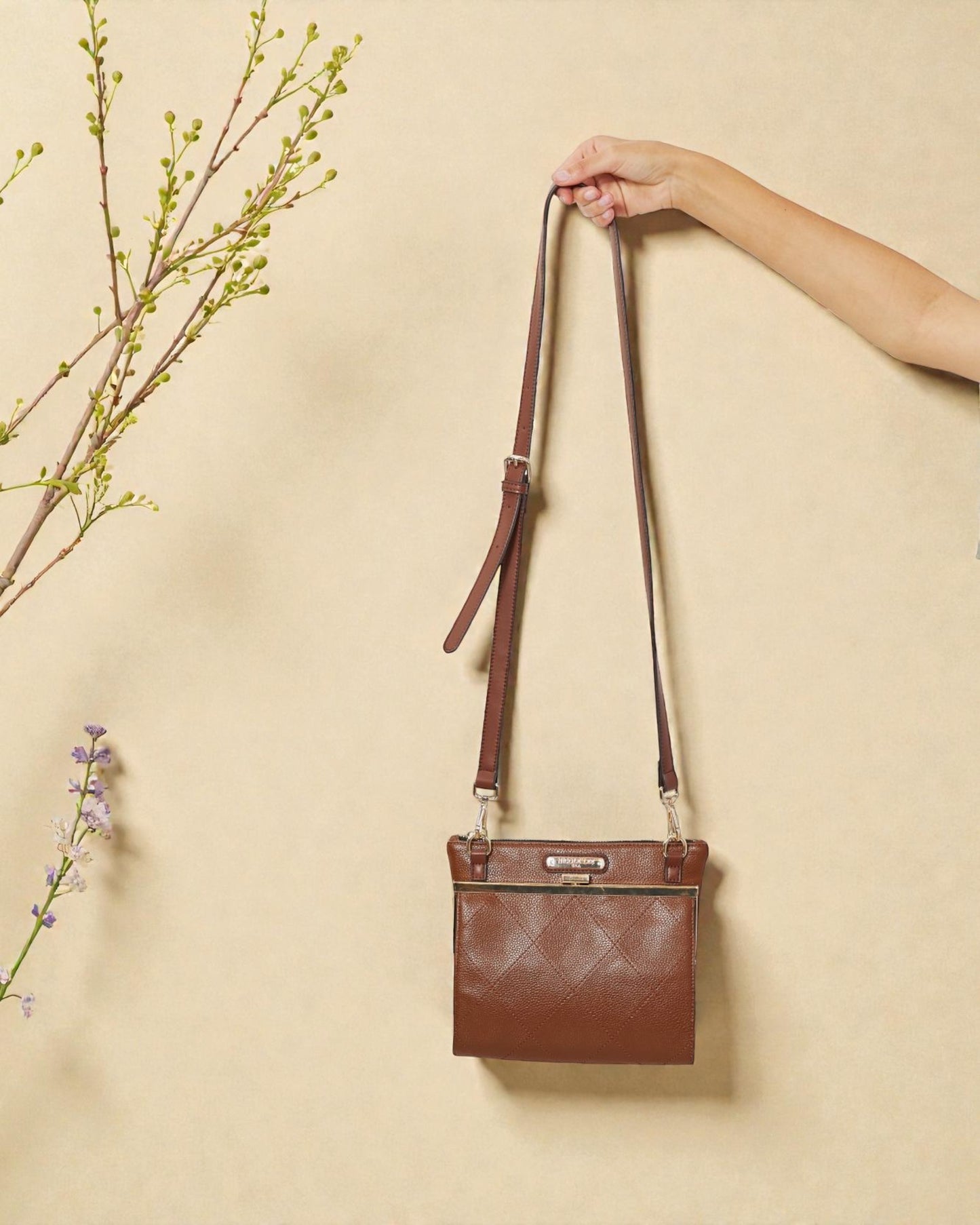 Brown leather crossbody bag held by a hand against a beige background with floral elements.