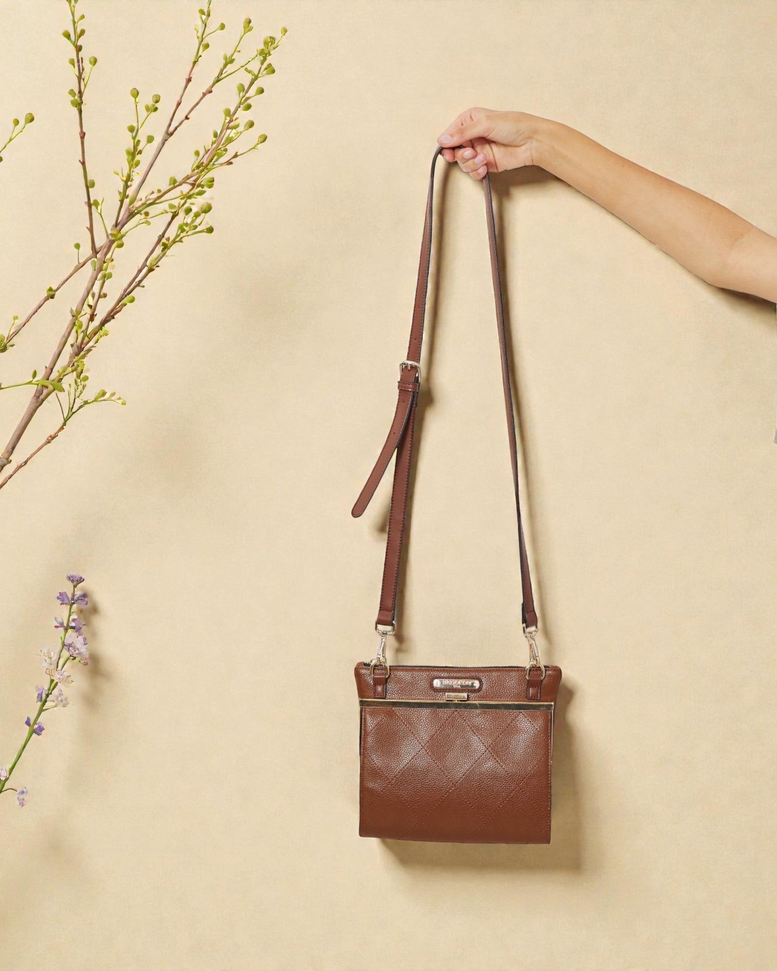 Brown leather crossbody bag held by a hand against a beige background with floral elements.