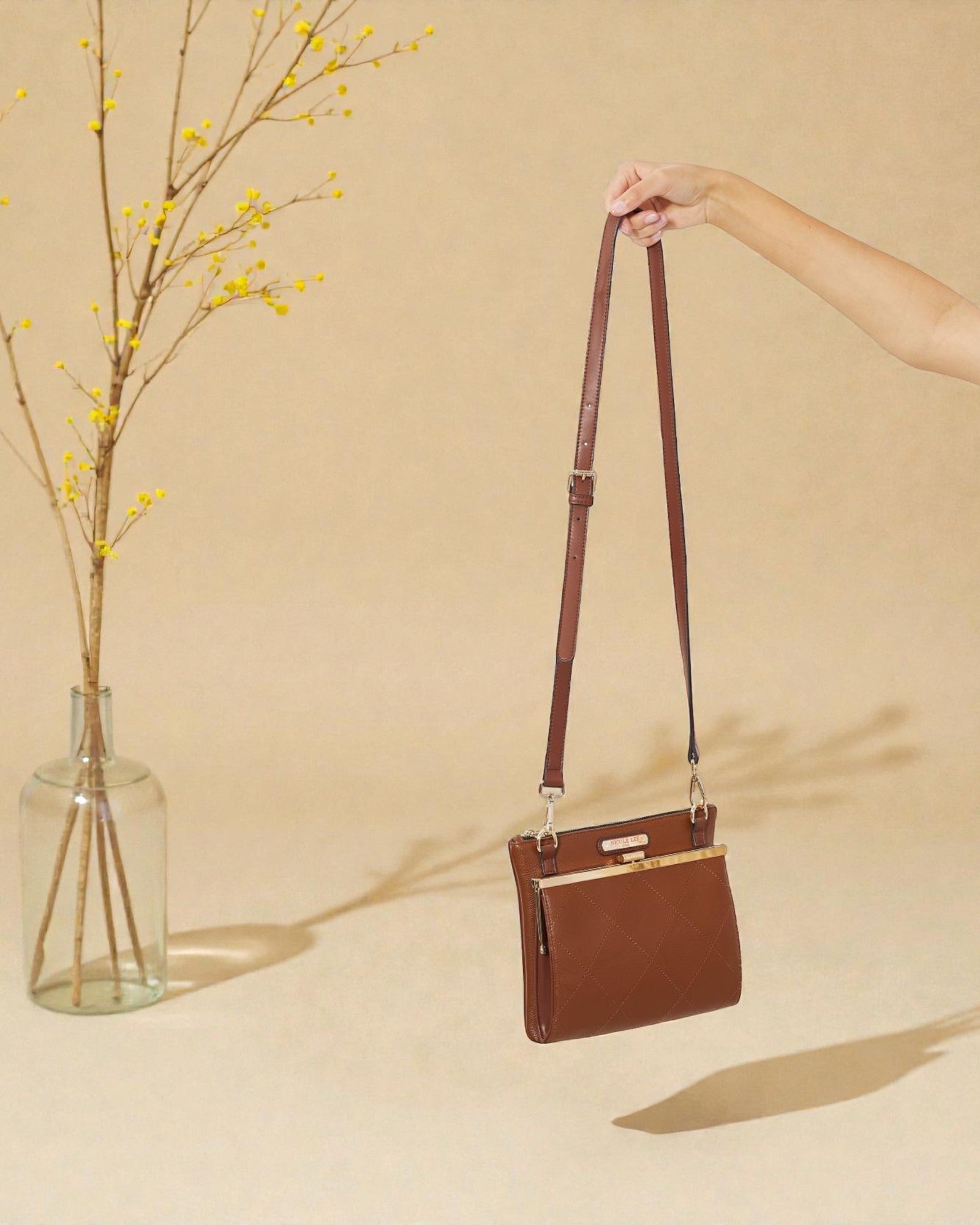 Brown handbag held by a hand against a beige background with a vase of yellow branches.