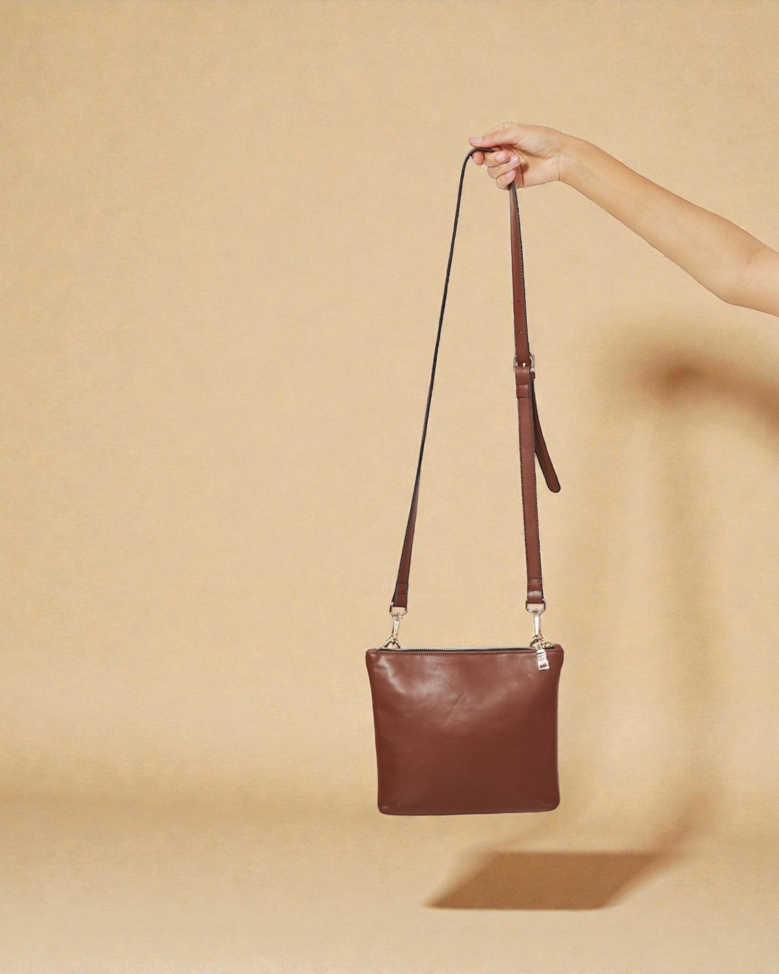 Brown leather handbag held by a hand against a beige background