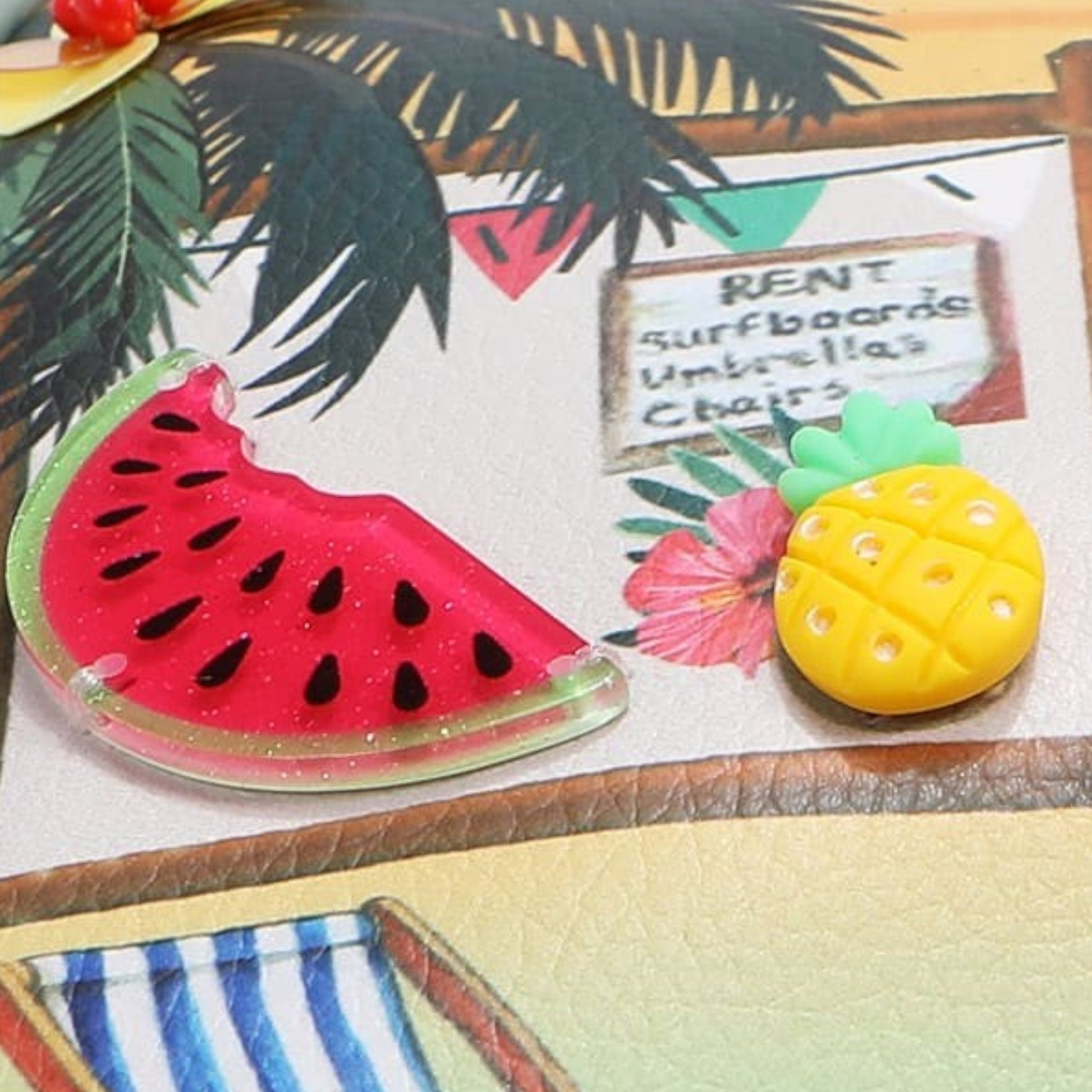 Two novelty pins, one shaped like a watermelon slice and the other like a pineapple, on a tropical-themed background.