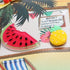 Two novelty pins, one shaped like a watermelon slice and the other like a pineapple, on a tropical-themed background.