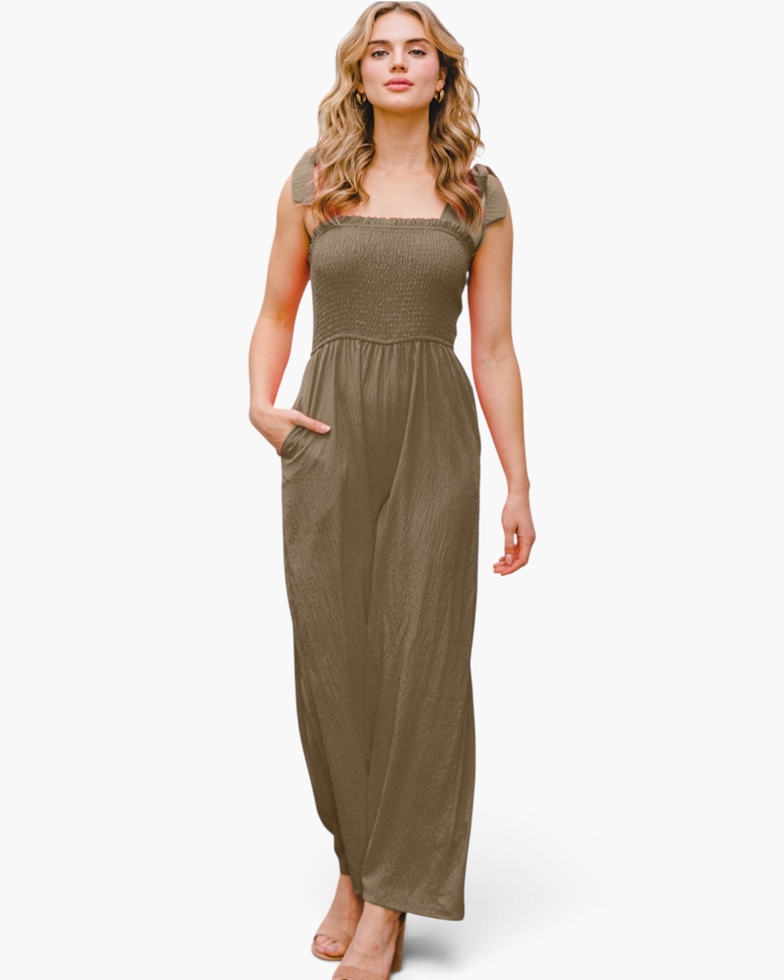 A woman modeling a sleeveless, smocked-bodice jumpsuit in a muted olive-brown color, featuring cap sleeves, a square neckline, and side pockets, set against a plain light gray background.