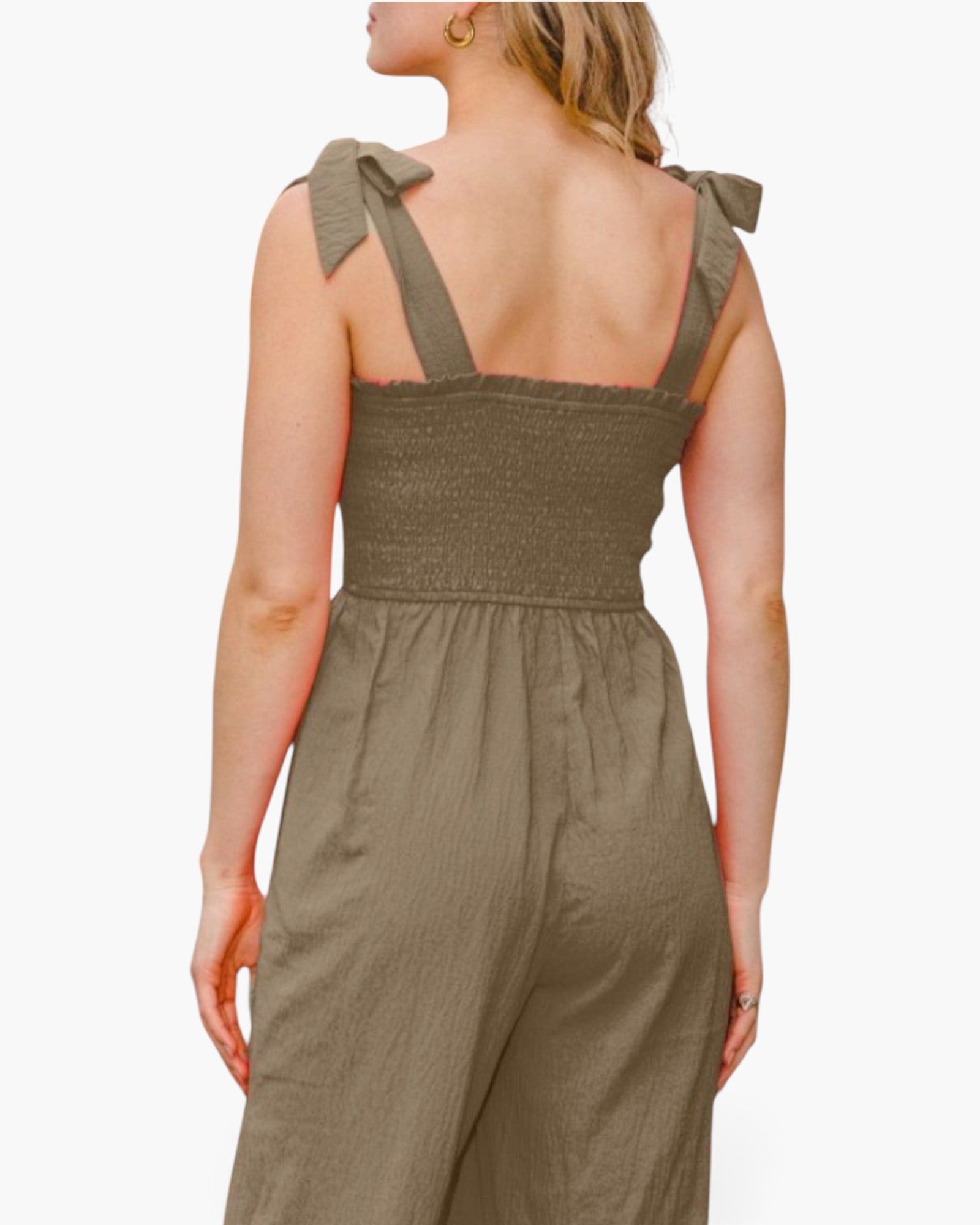 A woman shown from the back wearing an olive green jumpsuit with tie-shoulder straps and a smocked bodice, against a plain light background.