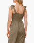 A woman shown from the back wearing an olive green jumpsuit with tie-shoulder straps and a smocked bodice, against a plain light background.