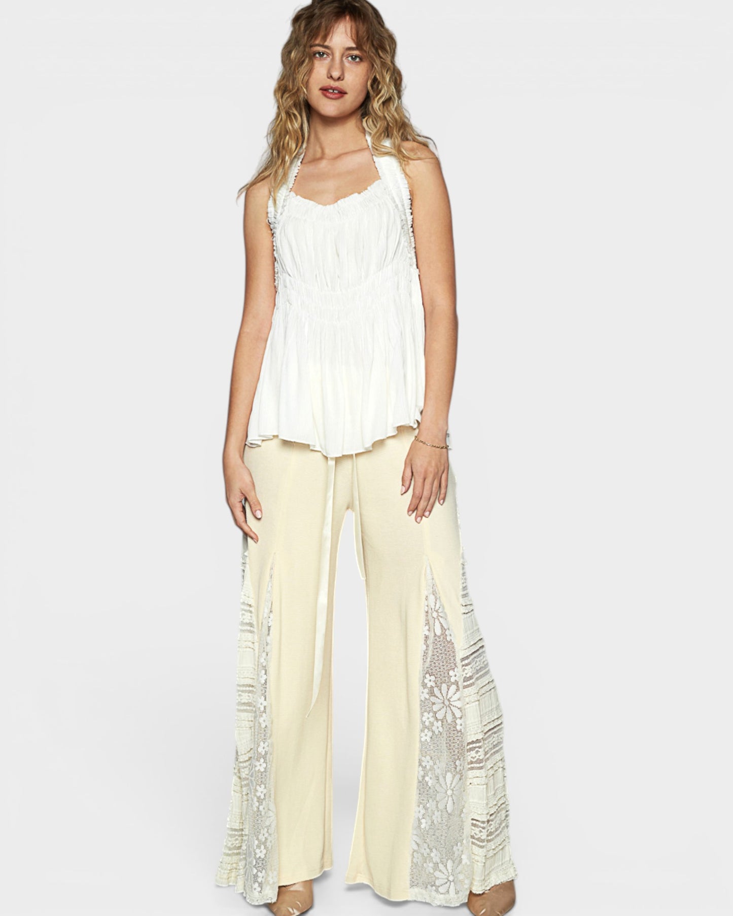 Woman wearing a white sleeveless top and cream pants with lace details on a light gray background
