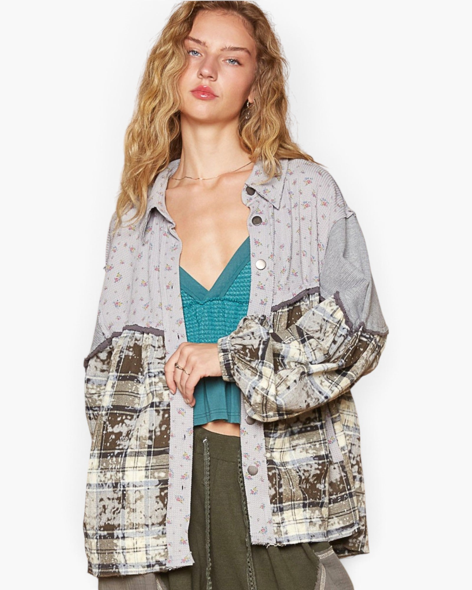 A woman models a mixed-media patchwork button-up shirt with a light gray base, floral print, and distressed plaid sleeves, layered over a teal knit top and olive green pants against a plain white background.