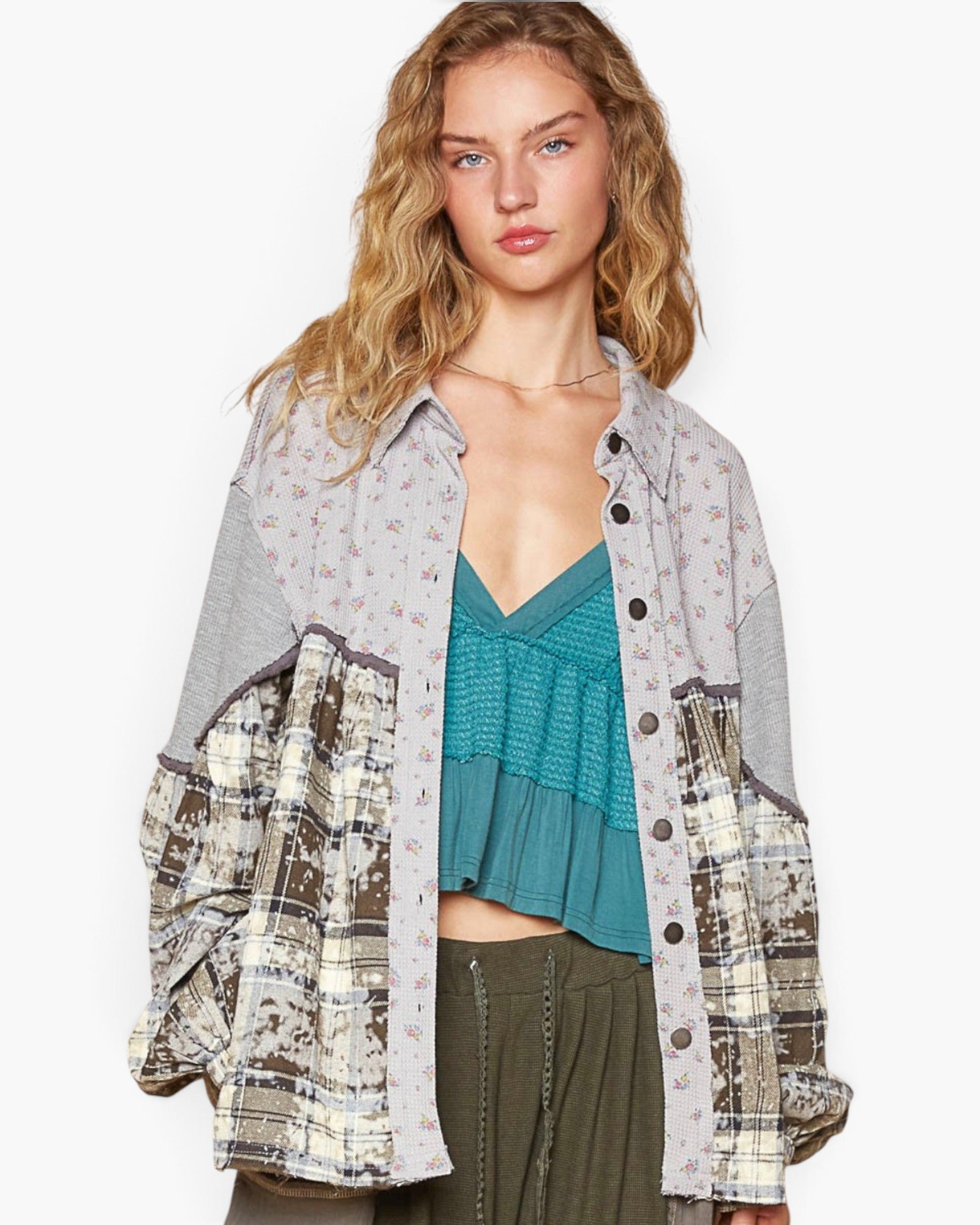 A woman modeling a patchwork button-up shirt with mixed prints including floral, plaid, and solid gray, worn over a teal top and olive green pants against a plain white background.