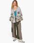 A model wearing a layered outfit featuring a light gray patterned button-up shirt with mixed prints, olive green wide-leg cargo pants, a teal crop top, and gray sneakers, set against a plain white studio background.