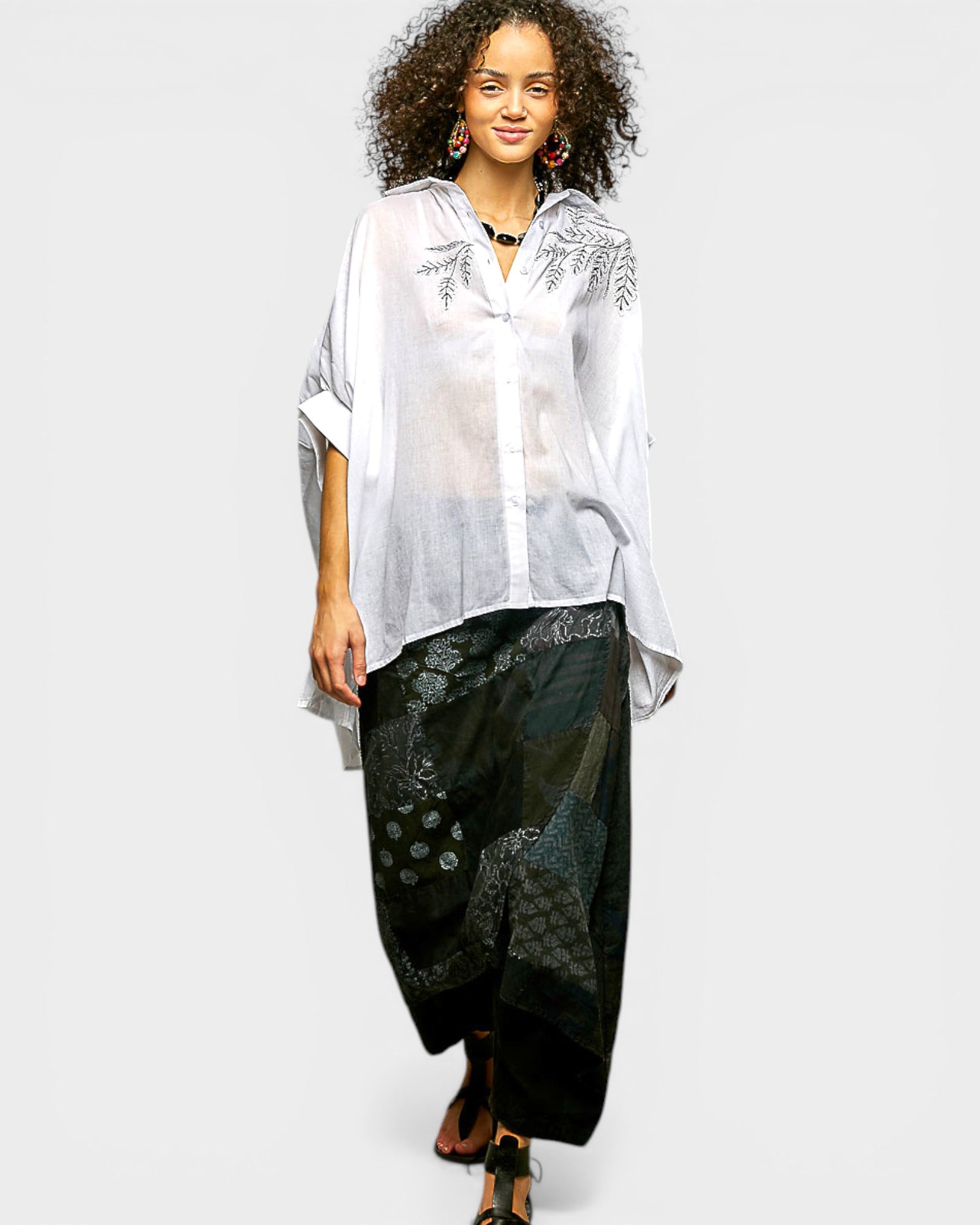 Woman wearing a white embroidered top and black patterned pants on a plain background