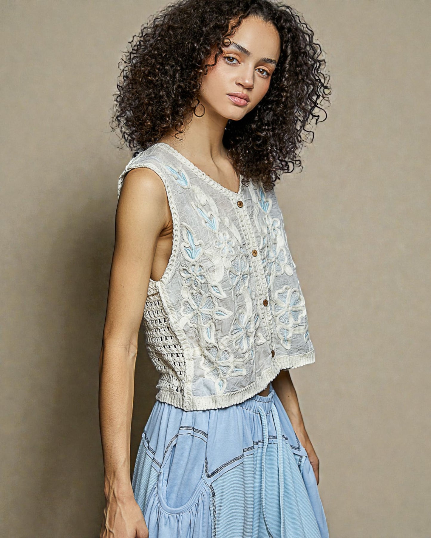 Woman wearing a sleeveless lace top and light blue pants against a beige background
