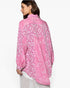 A woman shown from the back wearing a pink patterned button-up shirt with a high-low hem and white pleated trousers, set against a plain white studio background.