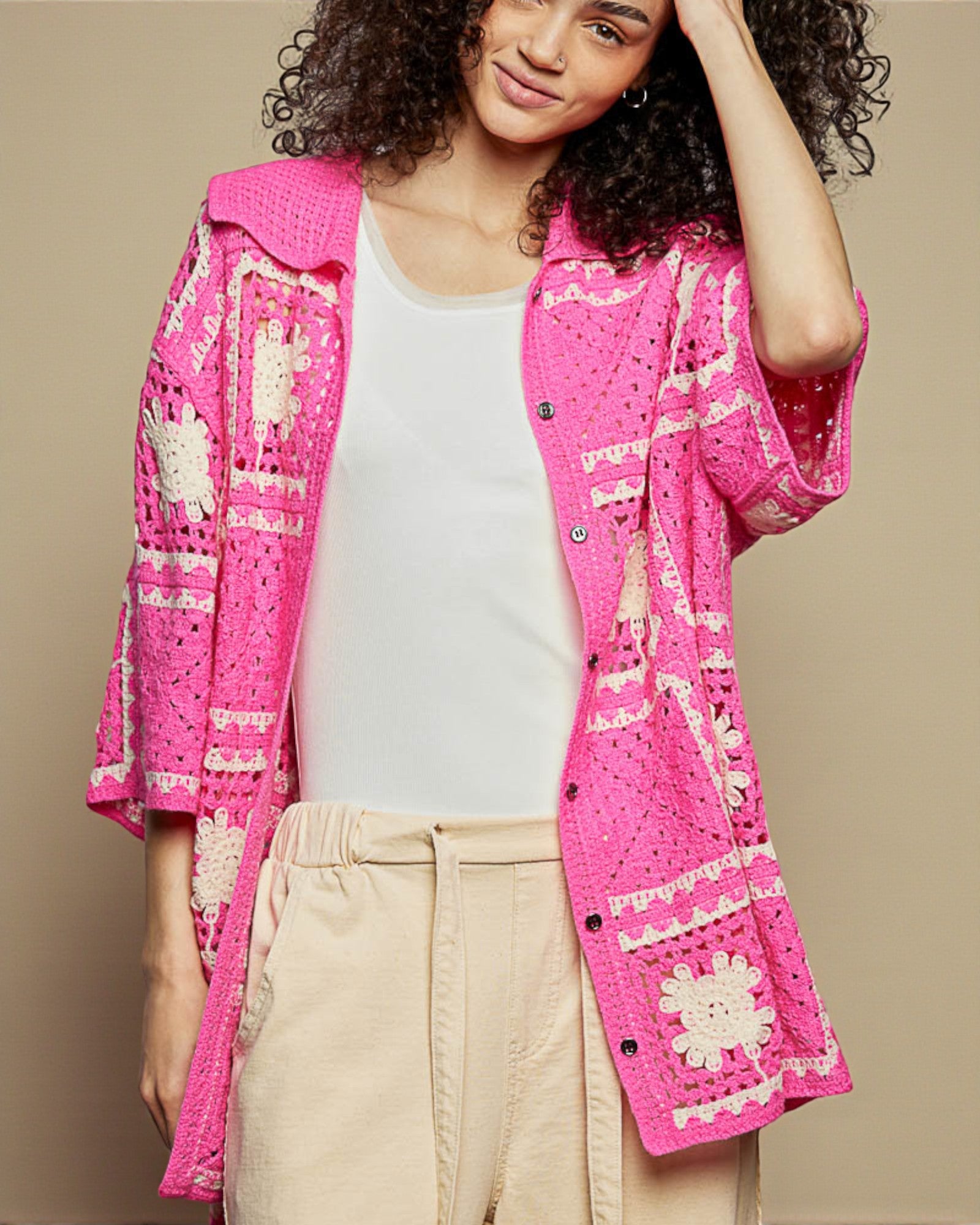 Woman wearing a bright pink crochet cardigan over a white shirt and beige pants on a beige background