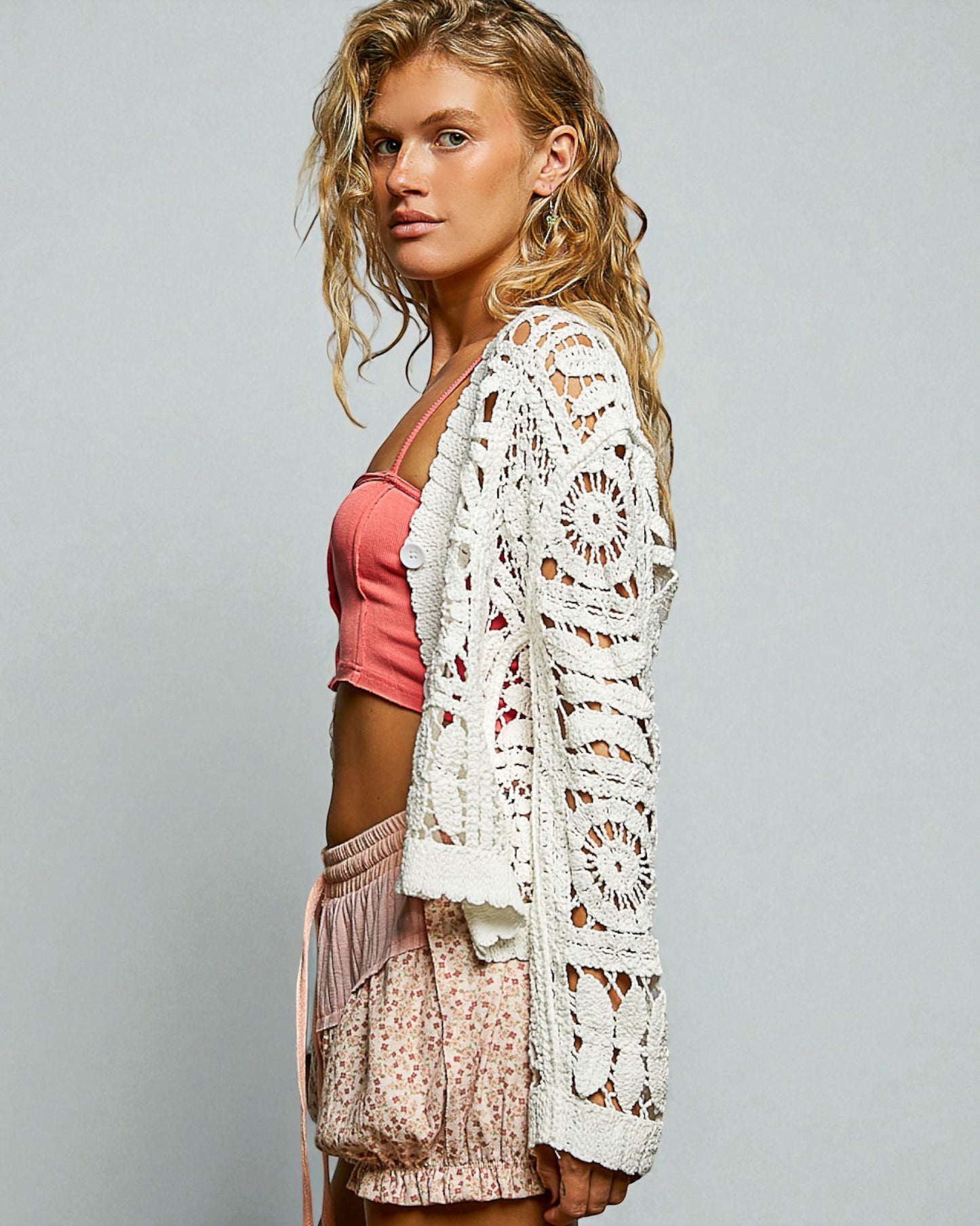Woman wearing a white crochet cardigan over a pink top and floral skirt on a gray background