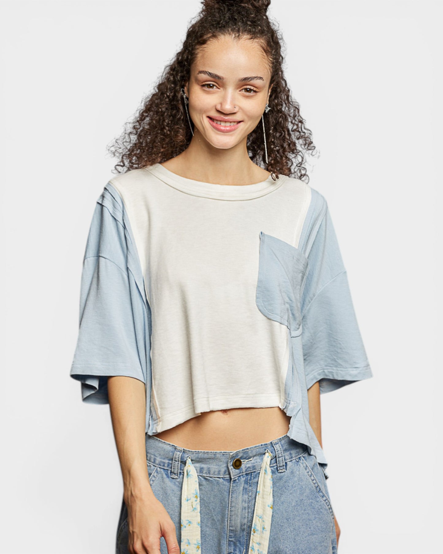 Woman wearing a white crop top with blue sleeves and denim pants on a light gray background