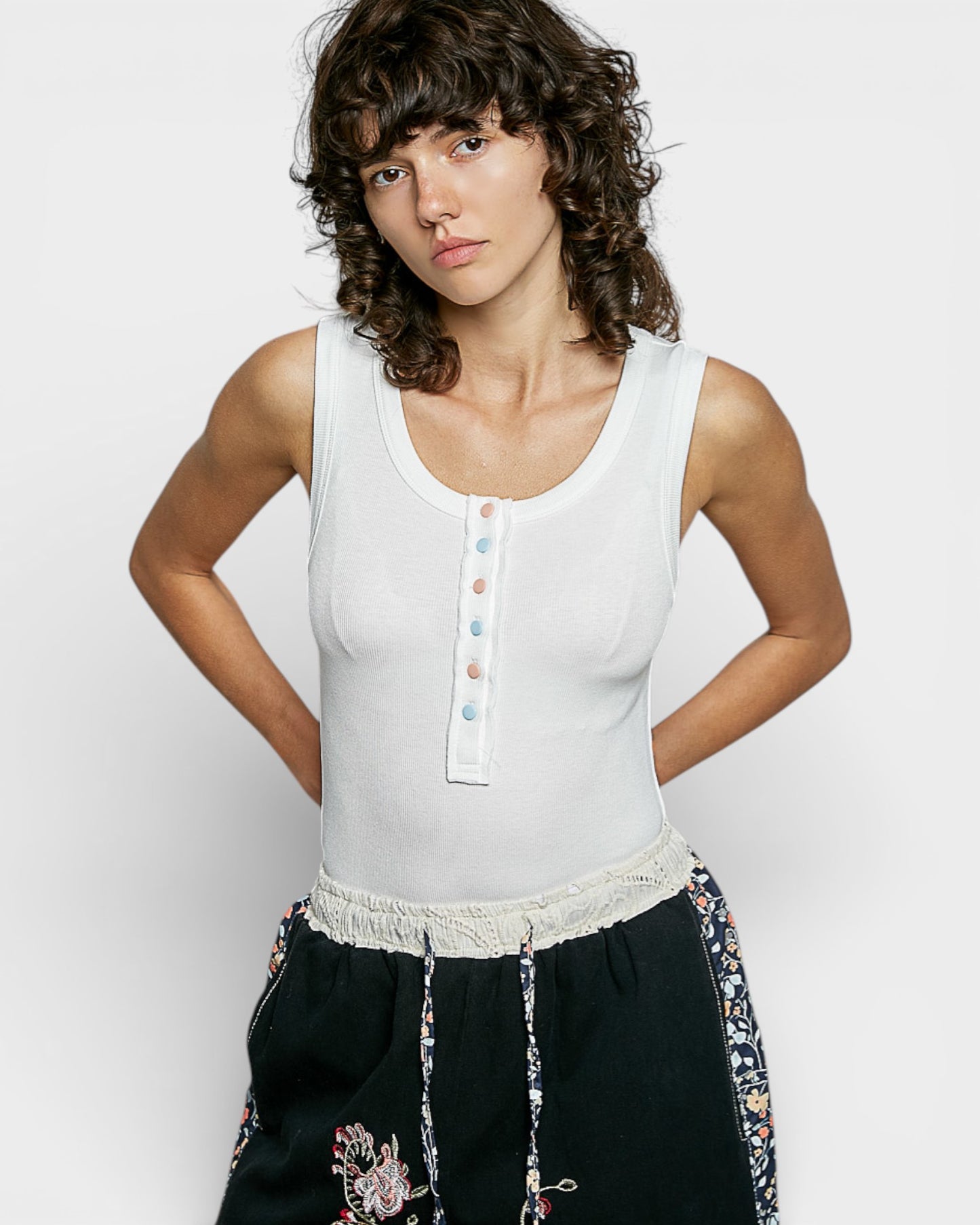 Woman wearing a white sleeveless top with colorful buttons and black pants with floral embroidery on a light gray background