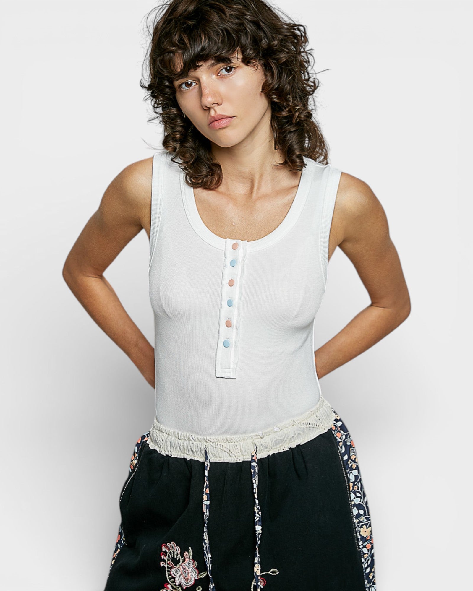 Woman wearing a white sleeveless top with colorful buttons and black pants with floral embroidery on a light gray background
