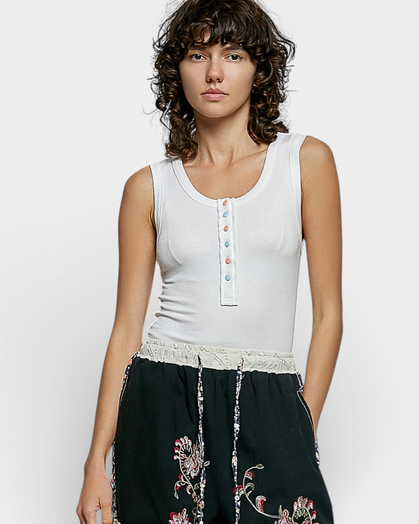 Woman wearing a white sleeveless top with colorful buttons and black pants with floral embroidery on a plain background
