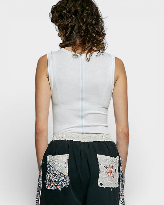 Person wearing a white sleeveless top and black pants with floral embroidery on a plain background