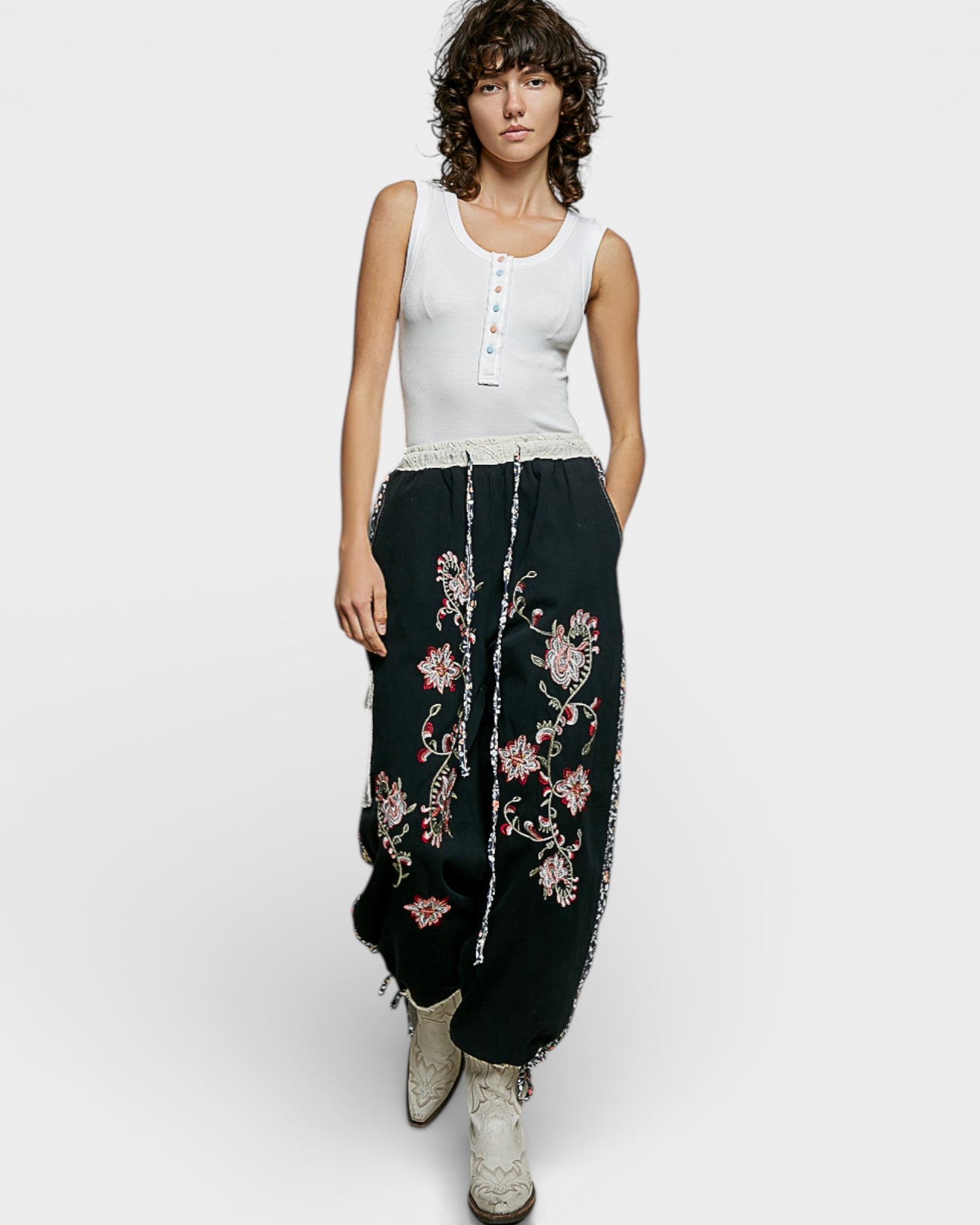 Woman wearing a white sleeveless top and black pants with floral embroidery on a white background