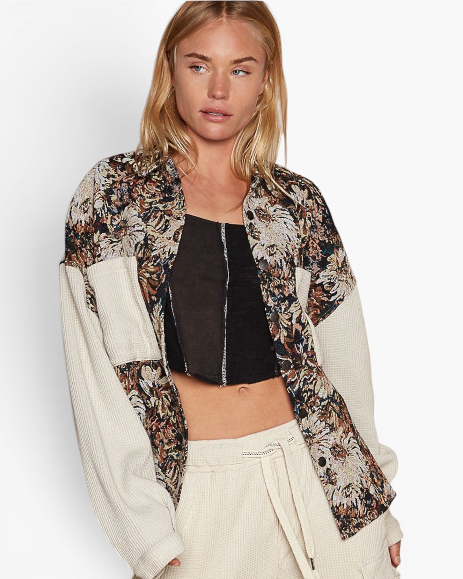 A woman modeling a color-blocked floral jacket with cream ribbed sleeves and a brown, beige, and black floral patterned body, worn over a black crop top and cream drawstring pants against a light gray studio background.