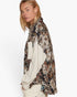 A woman shown in side profile wearing a long-sleeve button-up shirt with a dark floral and botanical print over a cream ribbed knit sweater, set against a plain white studio background.