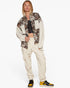 A model wearing a cropped jacket with a brown and beige floral pattern on the upper half and solid beige sleeves, paired with beige cargo pants and yellow and black sneakers, against a plain white background.