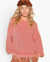 A woman wearing a coral pink ribbed knit sweater with a ruffled crew neck and distressed hem, paired with a brown patterned beret and brown pants, against a plain white background.