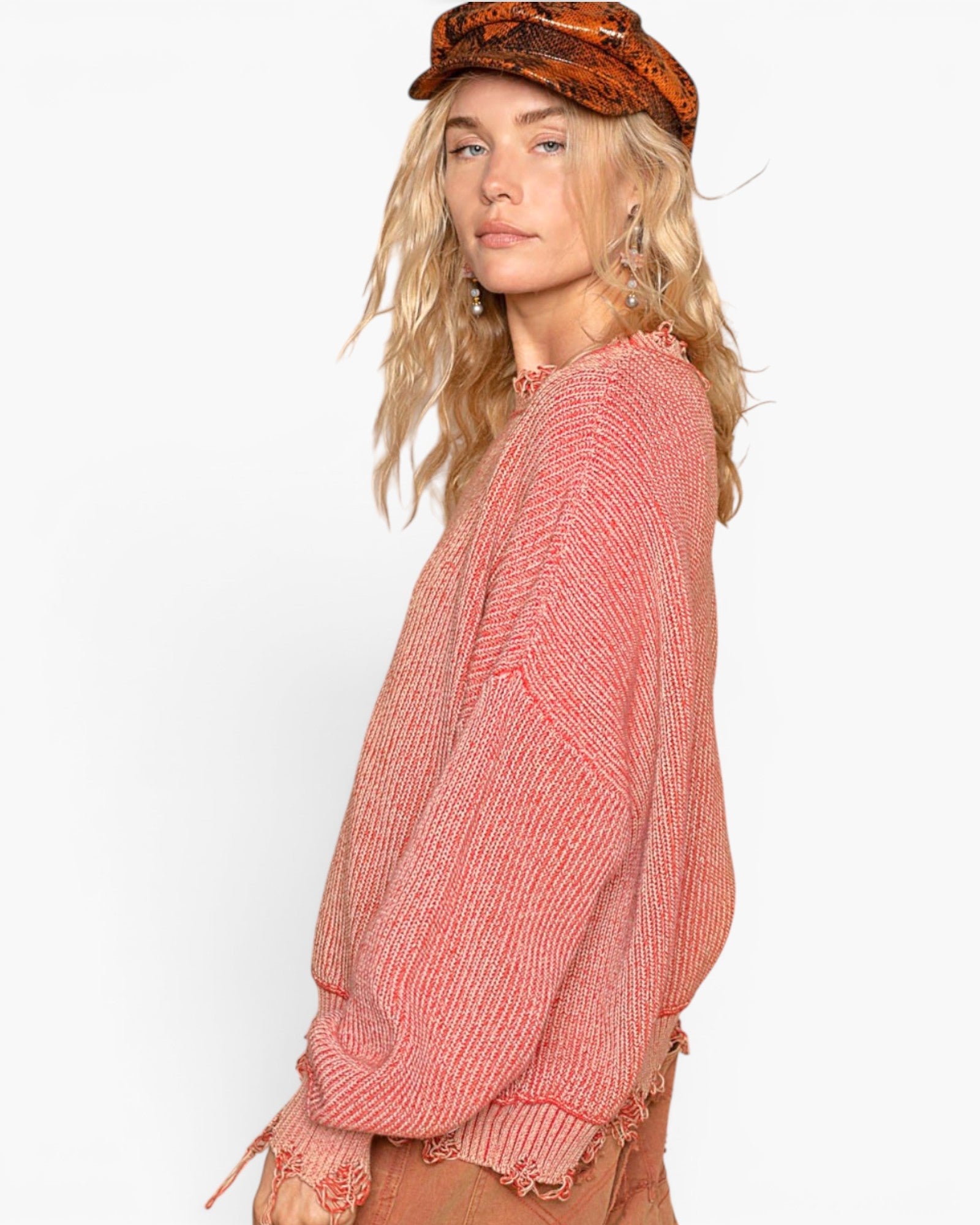 A woman wearing a textured, ribbed knit sweater in a dusty coral pink color, paired with a brown patterned beret and tan pants, photographed against a solid white studio background.