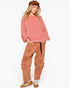 A model wearing a pink marled knit sweater, distressed rust-orange cargo pants, and brown sneakers, paired with a brown aviator hat, against a plain white background.