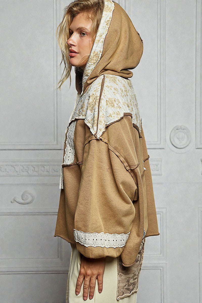 Woman wearing a beige hooded garment with lace details against a white wall.