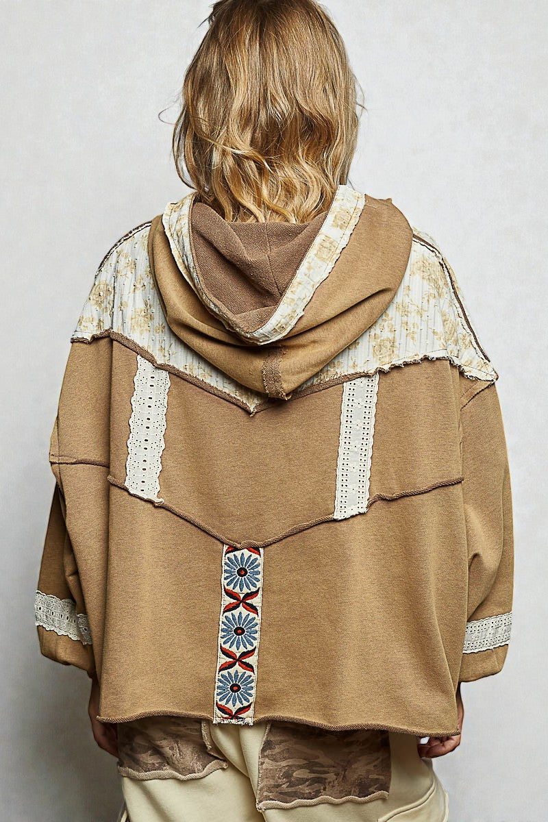 Person wearing a brown hoodie with decorative patterns on a plain background