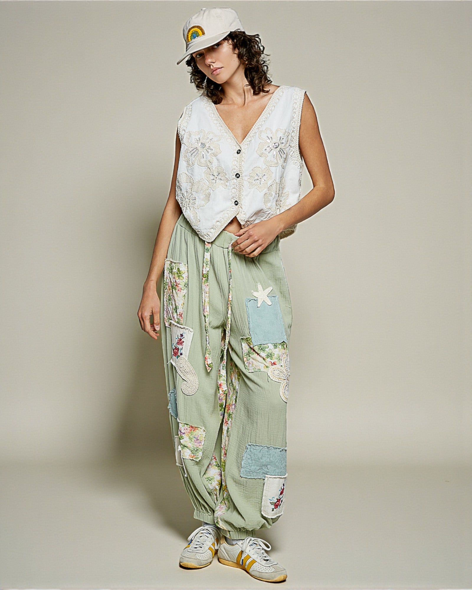 Woman wearing a white sleeveless top and green patchwork pants with a cap on a plain background