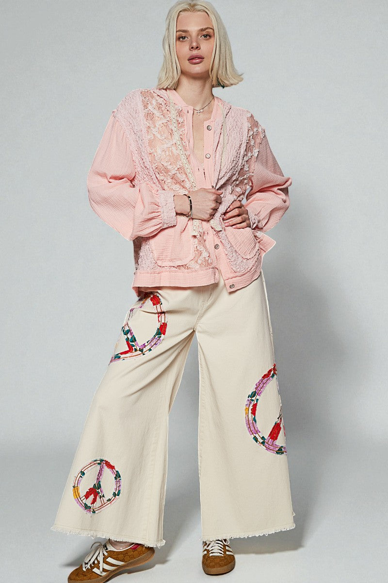 Woman wearing a pink embroidered blouse and cream pants with peace symbol designs on a plain background