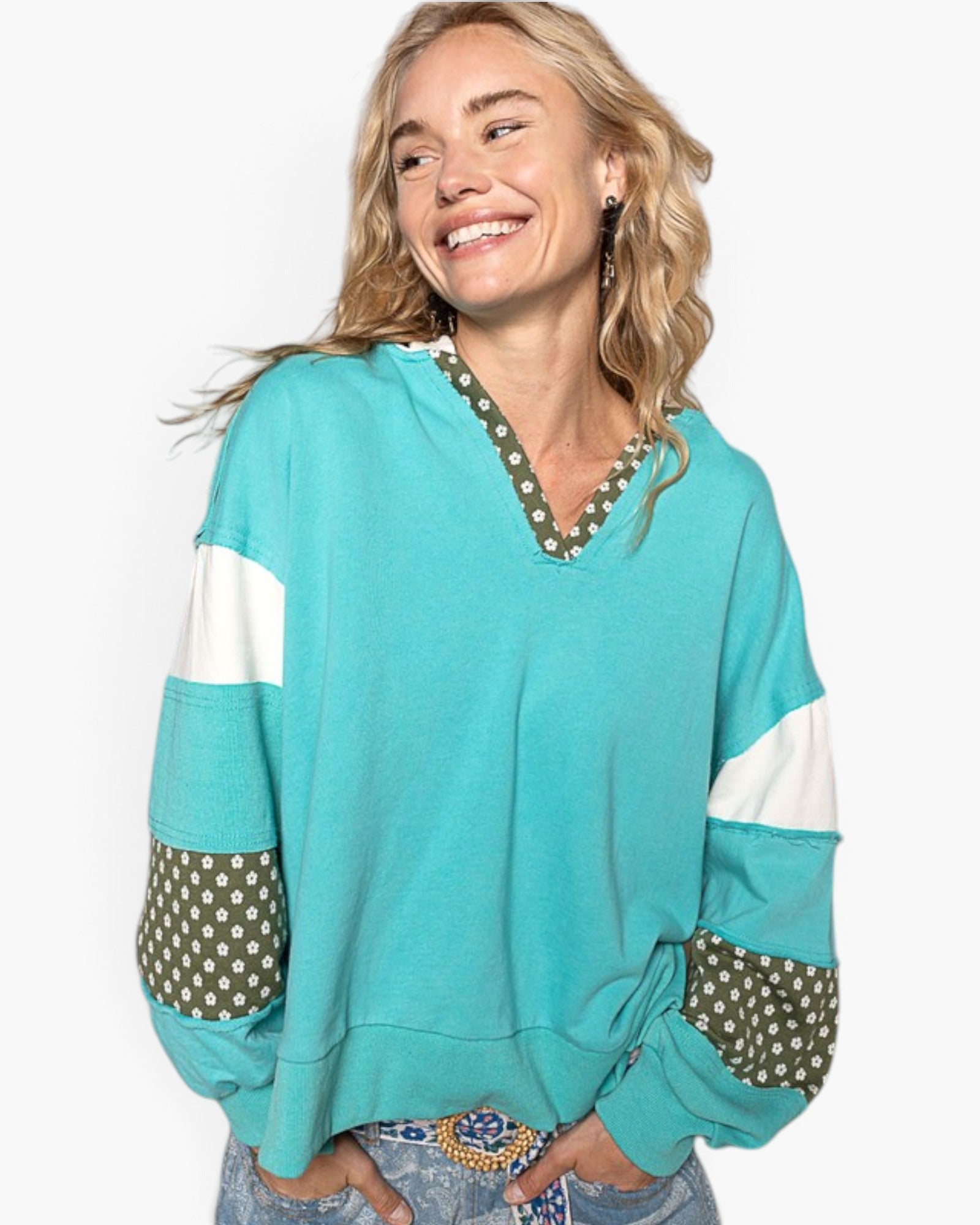 A woman wearing a turquoise color-blocked pullover sweatshirt with white sleeve stripes and olive green polka-dot patchwork details on the cuffs and V-neckline, paired with blue jeans against a plain white background.