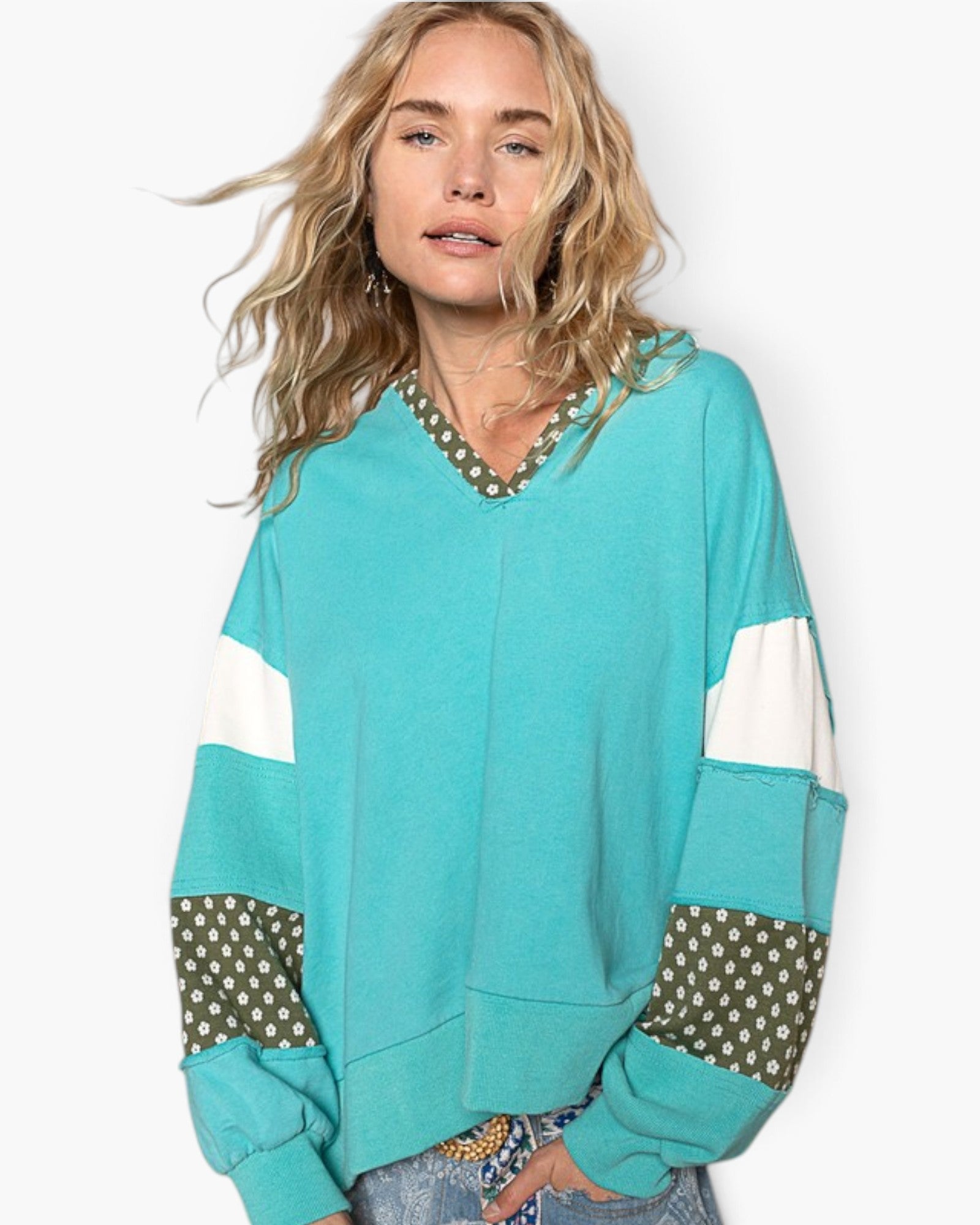 A woman wearing a turquoise color-block sweatshirt with white panels and olive green polka-dot patterned accents on the neckline and cuffs, paired with blue jeans against a light gray studio background.